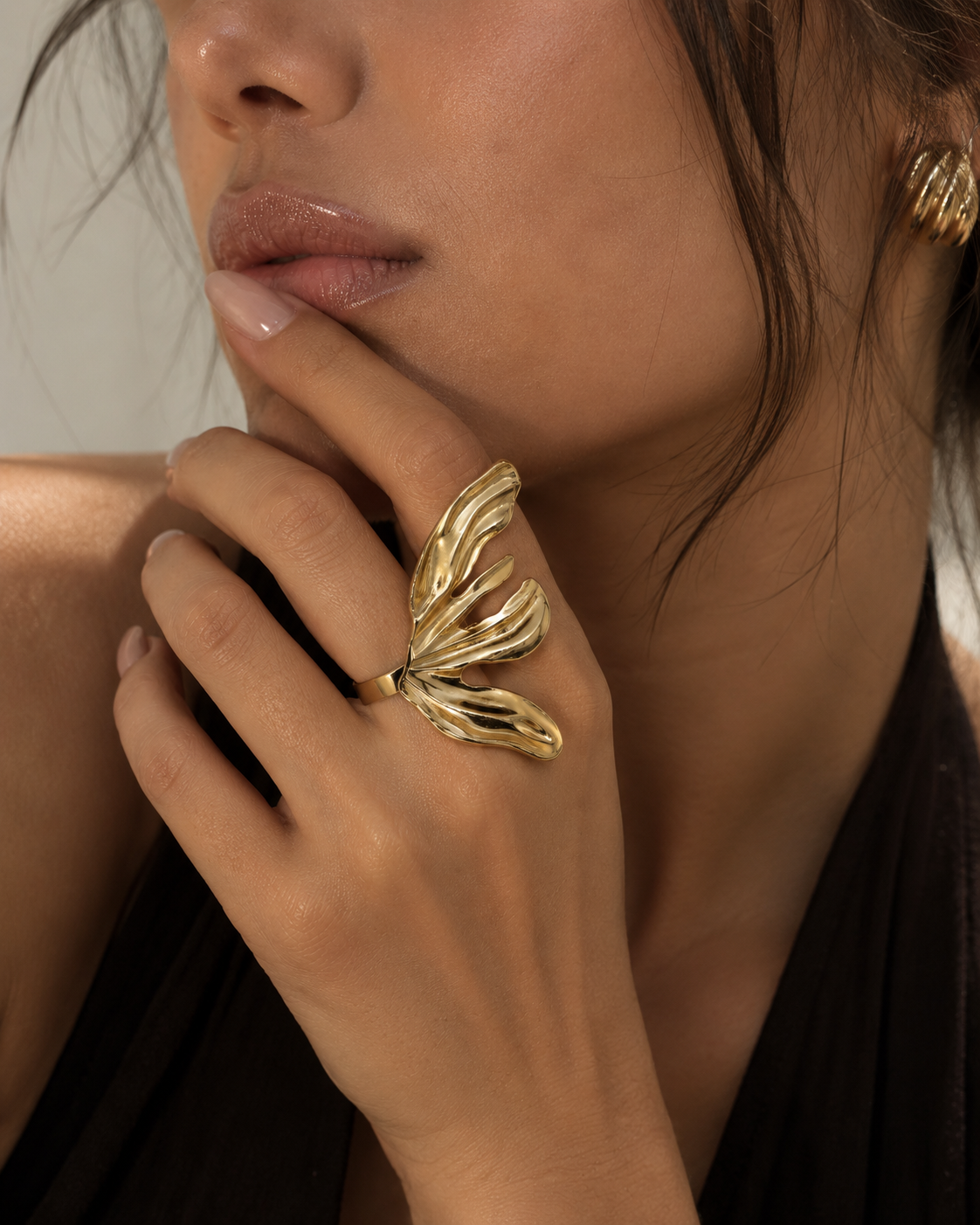 A close-up lifestyle shot of a model wearing a chunky, organic-shaped gold ring on her finger, with a matching gold earring visible on her ear.