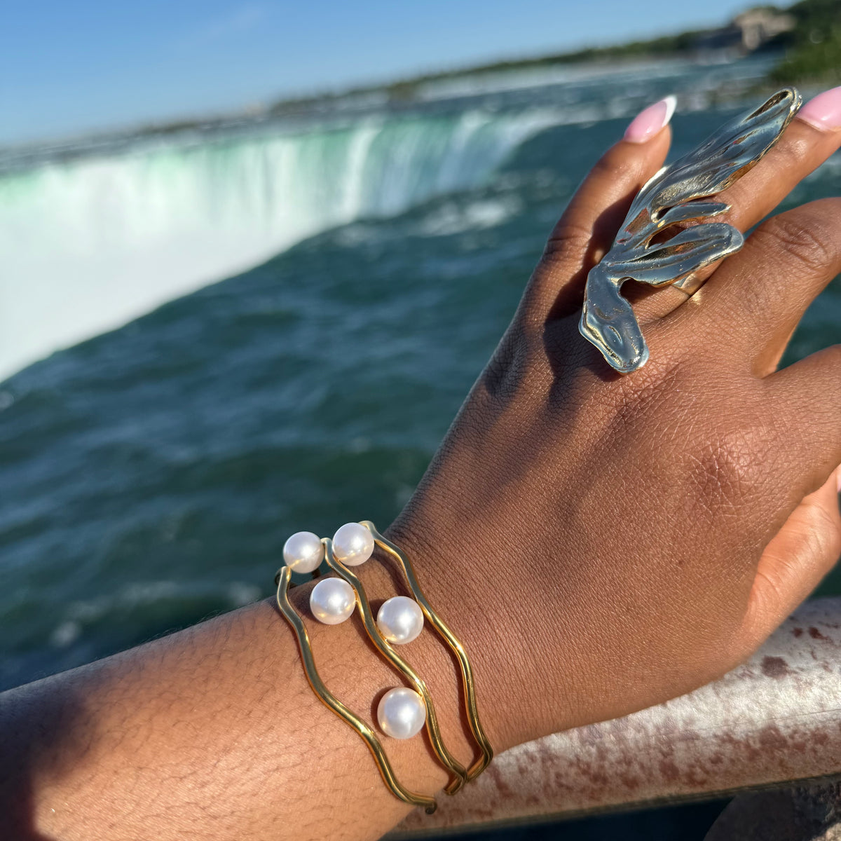 Hand wearing a gold bracelet with pearls and a silver ring, with a blurred waterfall background
