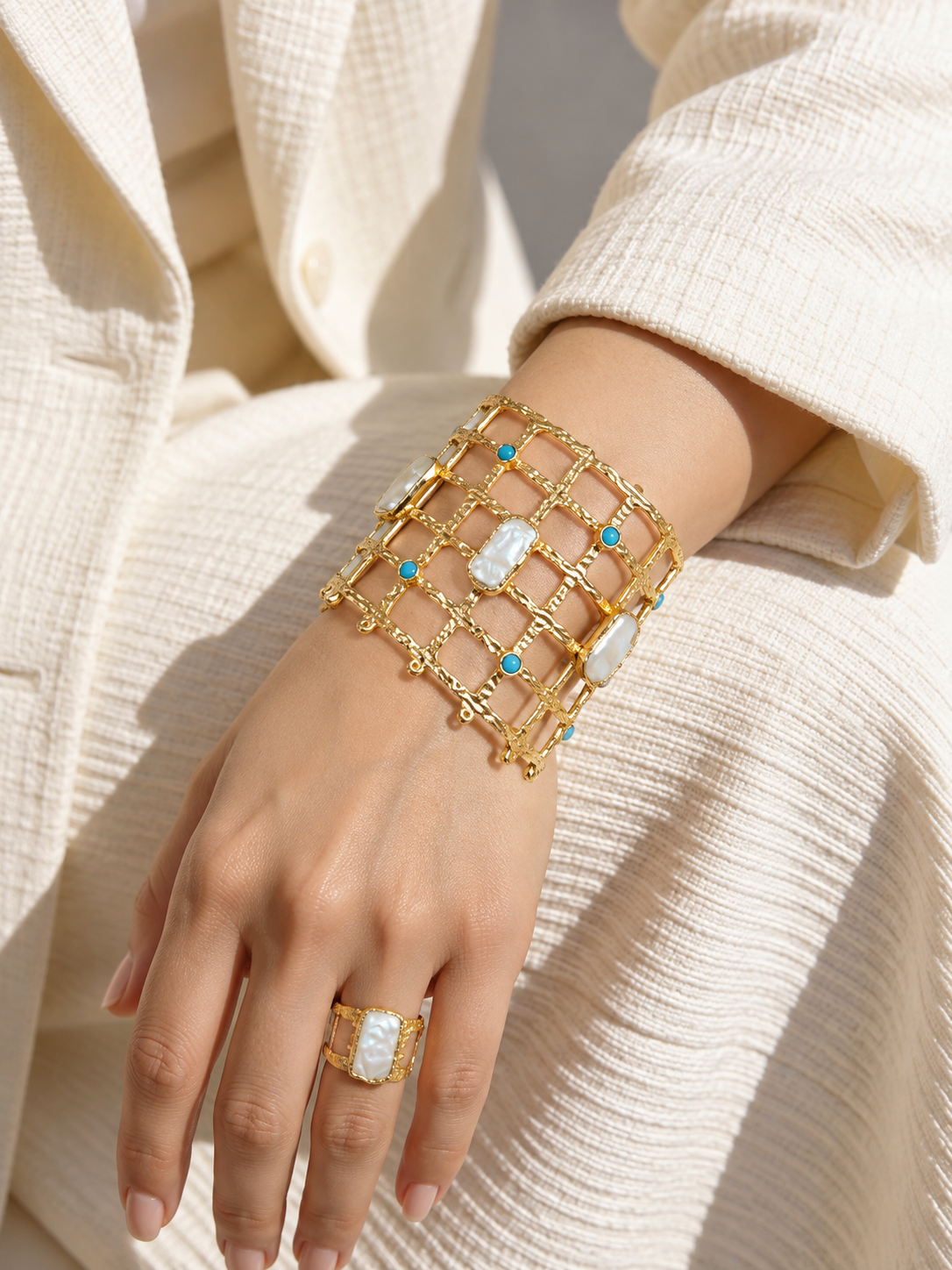 A close-up of a person's hand and wrist wearing a wide gold lattice bracelet with white mother-of-pearl rectangular inlays and small turquoise blue stones, paired with a matching gold ring.