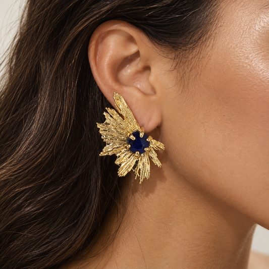 A close-up of a model's ear wearing a textured gold leafburst earring with a deep blue gemstone at the center.