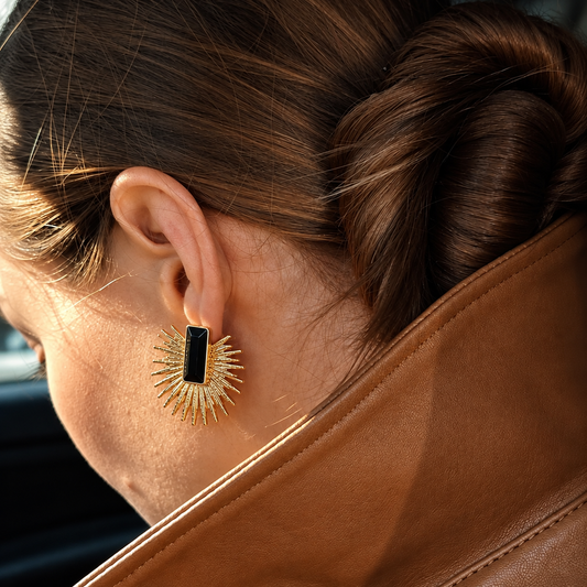 A close-up lifestyle shot of a woman wearing a gold sunburst earring with a black rectangular center, set against her brown hair and tan leather jacket.