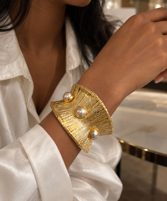 A close-up of a person wearing a wide textured gold cuff bracelet with three white pearl accents, paired with a white satin blouse.