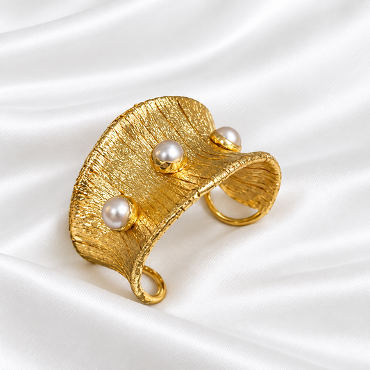 A textured gold cuff bracelet with three white faux-pearl accents, displayed on a draped white satin fabric background.