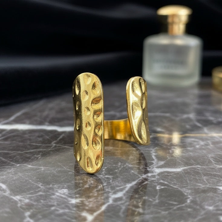 Gold ring with textured design on a marble surface with a blurred perfume bottle in the background.