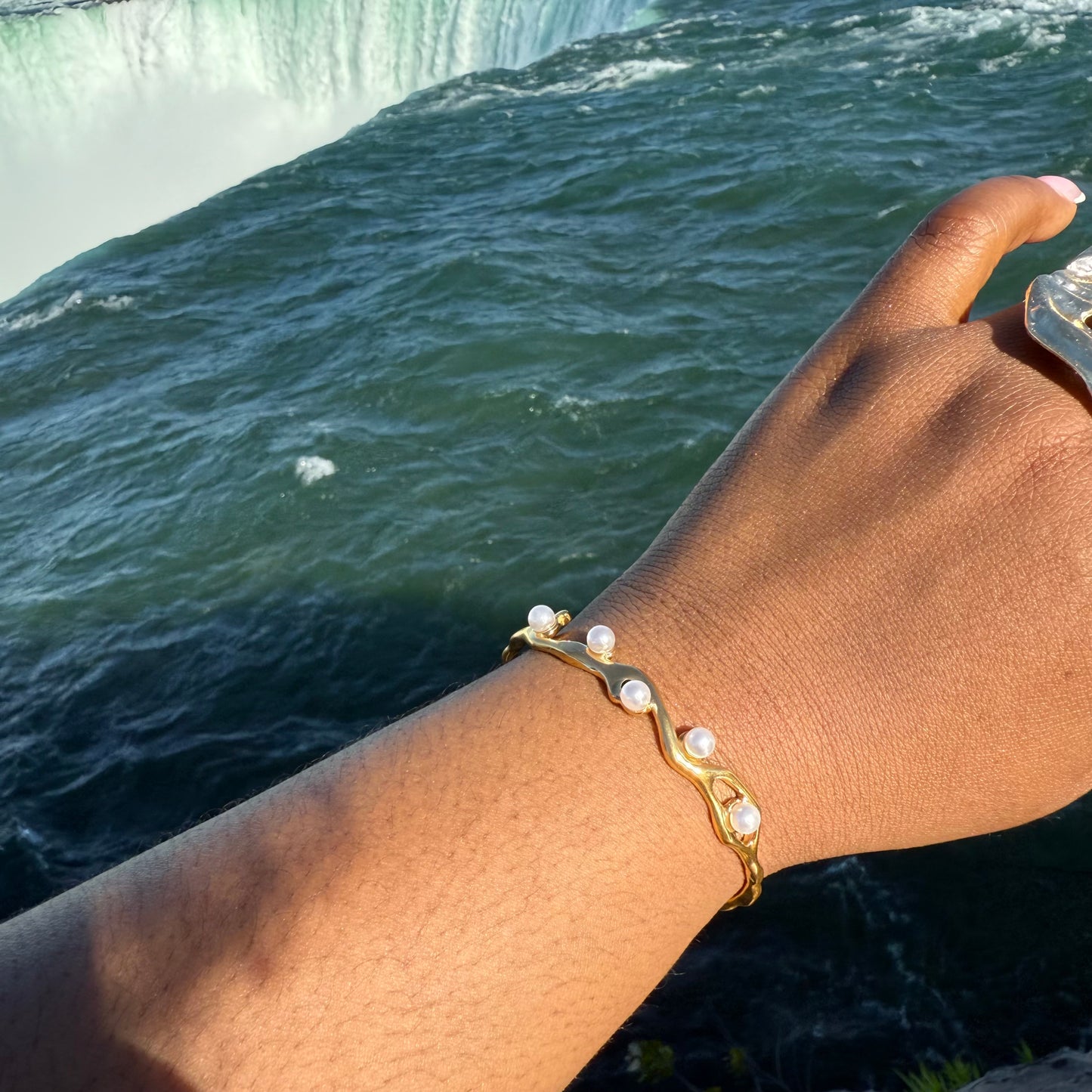 Gold bracelet with pearls on a wrist against a backdrop of a waterfall.