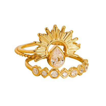 A gold ring featuring a sunburst design with a teardrop-shaped clear gemstone in the center and a lower band adorned with small round clear gemstones, shown on a plain white background.