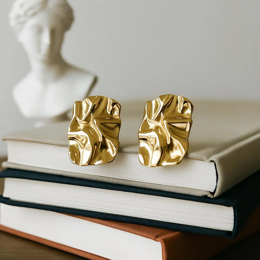 A pair of chunky, textured gold stud earrings with a crumpled, organic surface, displayed on a stack of books with a white classical bust statue in the blurred background.