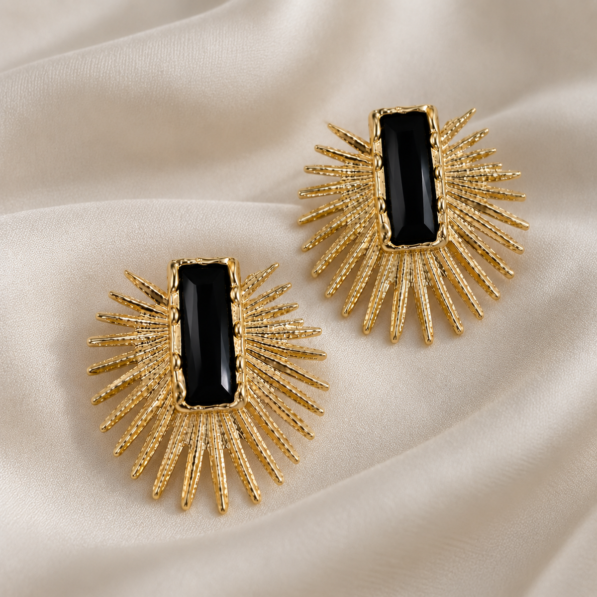 A pair of gold-toned statement earrings with a rectangular black stone in the center and radiating textured gold spikes, displayed on a draped cream satin fabric.