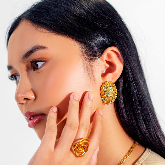 Asian Woman wearing herae gold earrings and ring, posing with hand by ear against a white background
