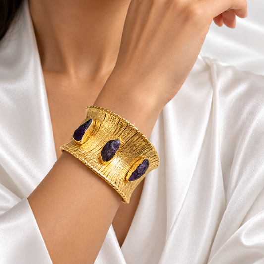 A close-up of a person's wrist wearing a wide textured gold cuff bracelet with three dark purple stone accents, set against a white satin robe.