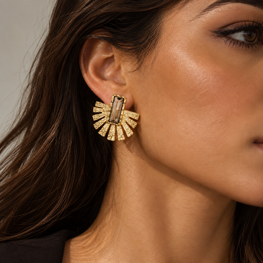 A close-up of a woman's ear wearing a gold fan-shaped stud earring with a rectangular smoky brown center stone and small clear stones around it.