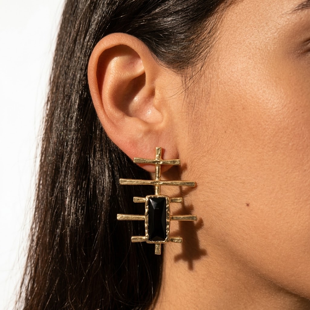 A close-up of a model's ear wearing a geometric gold earring with a rectangular black stone.