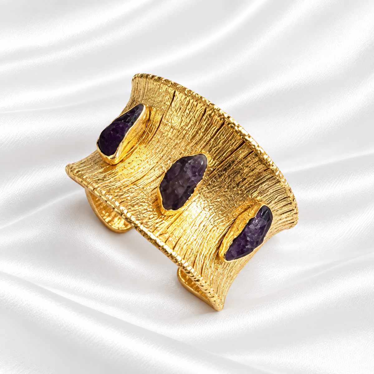 A wide gold cuff bracelet with a textured, brushed finish and three dark purple raw stone inlays, displayed on a white satin fabric background.