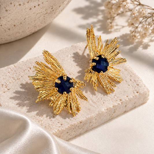 A pair of gold-toned statement earrings with textured sunburst petals and deep blue faceted centers, displayed on a light stone slab with soft fabric and dried flowers in the background.