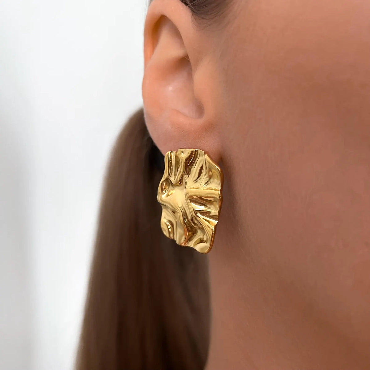 A close-up of a model's ear wearing a textured, organic-shaped gold earring.