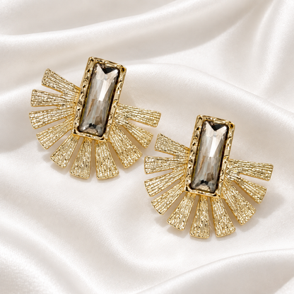 A pair of gold-toned statement earrings with a rectangular clear faceted gemstone in the center and textured fan-like rays extending outward, displayed on a white satin fabric background.