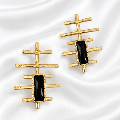 A pair of gold geometric stud earrings with rectangular black inlays, displayed on a white satin fabric background.