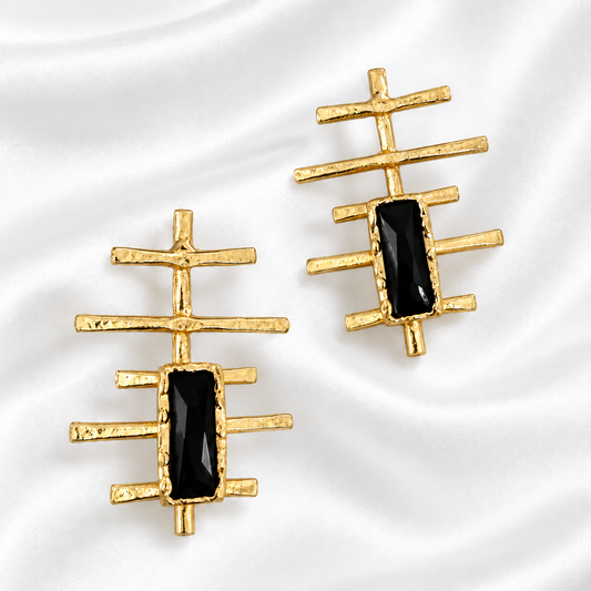 A pair of gold geometric stud earrings with rectangular black inlays, displayed on a white satin fabric background.
